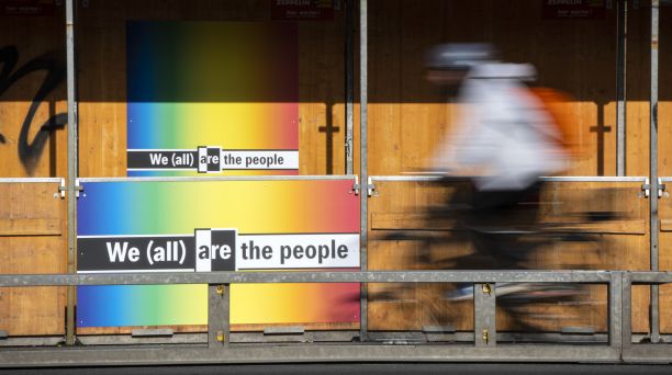 Bauzaun des Neubaus für die Neue Nationalgalerie. Auf zwei Plakaten steht auf regenbogenfarbigem Hintergrund in schwarz-weiß geschrieben „We (all) are the people“.