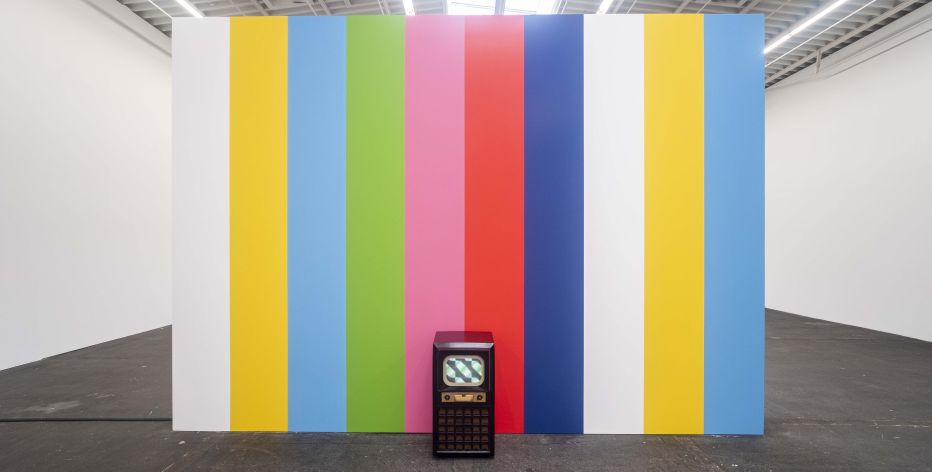 Vor einer Wand mit bunten Streifen, die an ein Fernsehtestbild von früher erinnern, steht ein altes Fernsehgerät mit Holzrahmen.